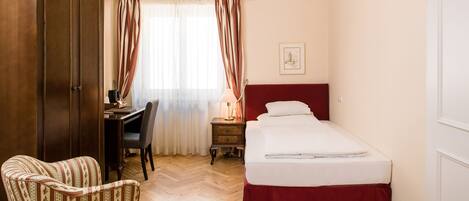 A hotel room with a wooden wardrobe, a desk with a chair, a bed with a red headboard, and a chandelier.