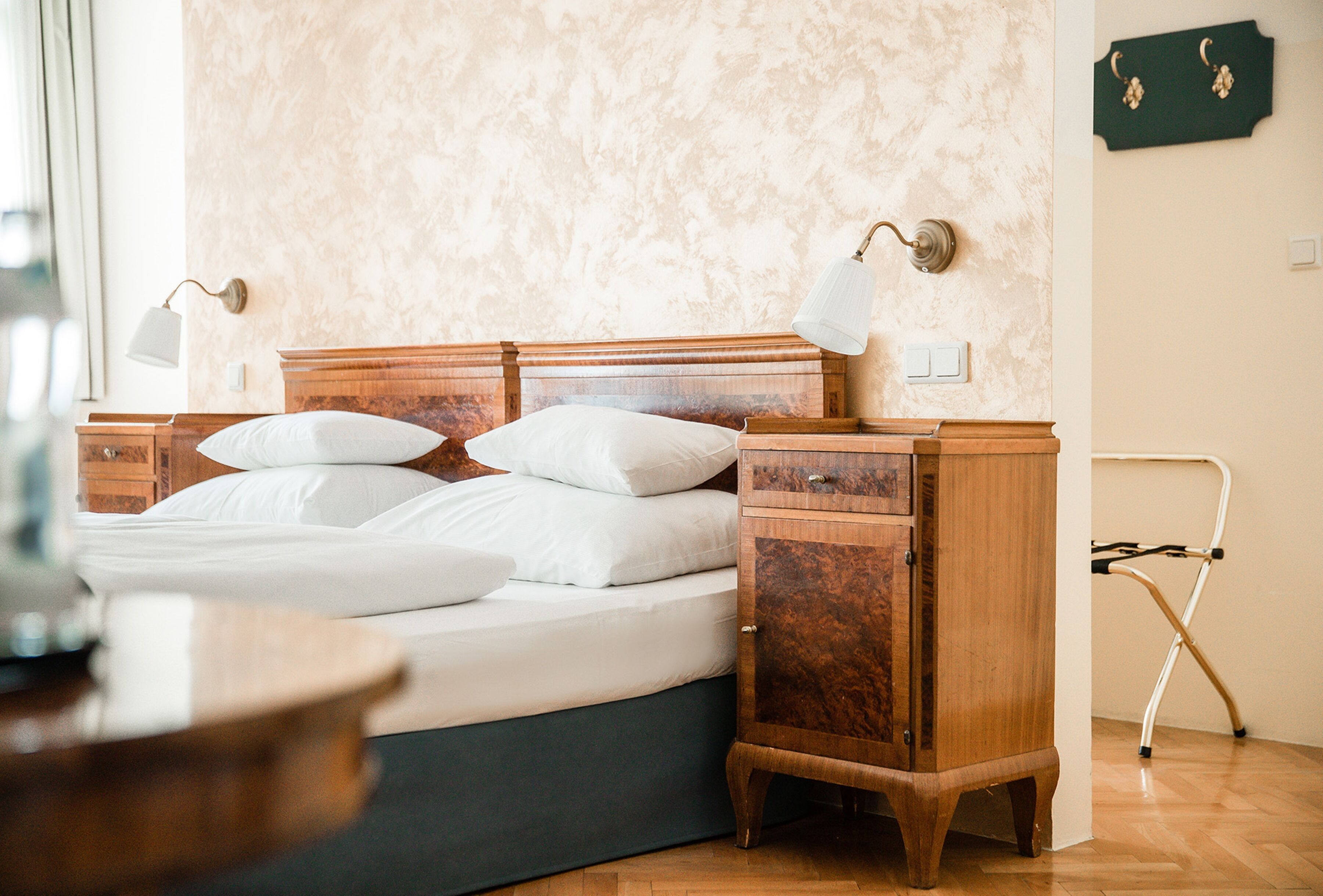 A neatly made bed with white linens, a wooden nightstand with a lamp, and a small folding chair.
