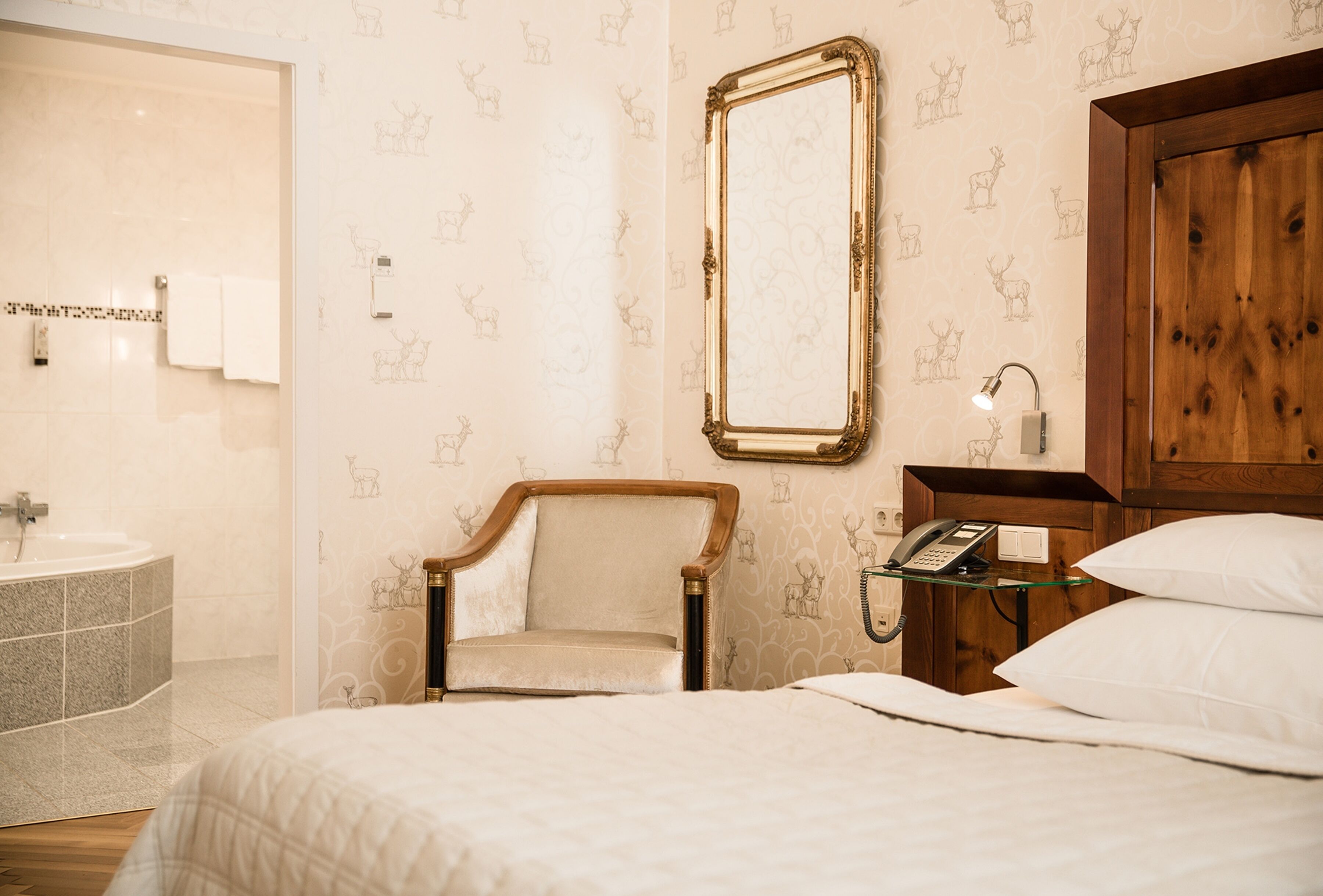 Une chambre d’hôtel comprenant un lit, une chaise, une armoire en bois, une table de chevet avec une lampe, et une salle de bain avec un lavabo et une baignoire.