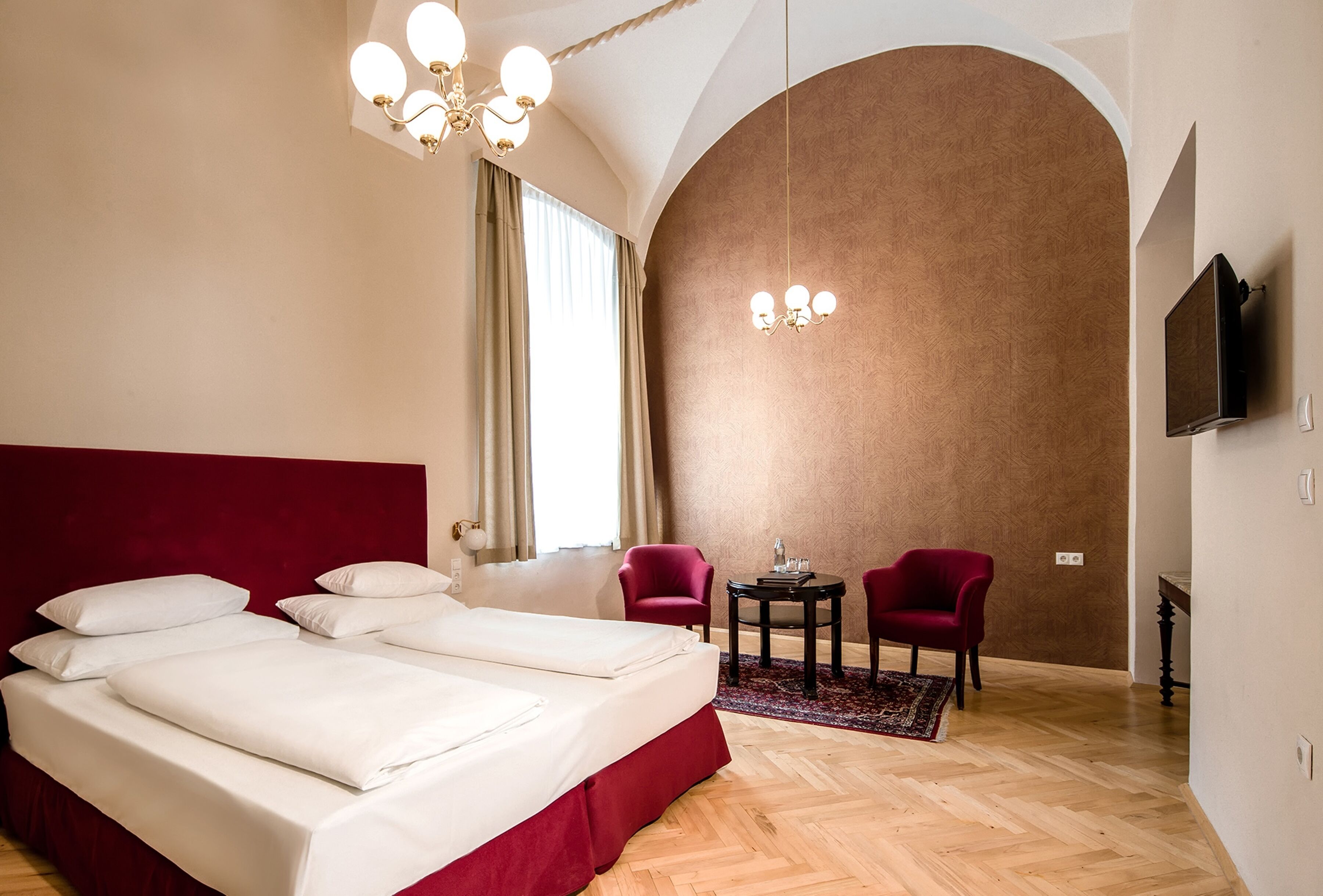Ein Hotelzimmer mit einem großen Bett, zwei roten Sesseln, einem kleinen Tisch, einem Fernseher und einer Chandelüster.