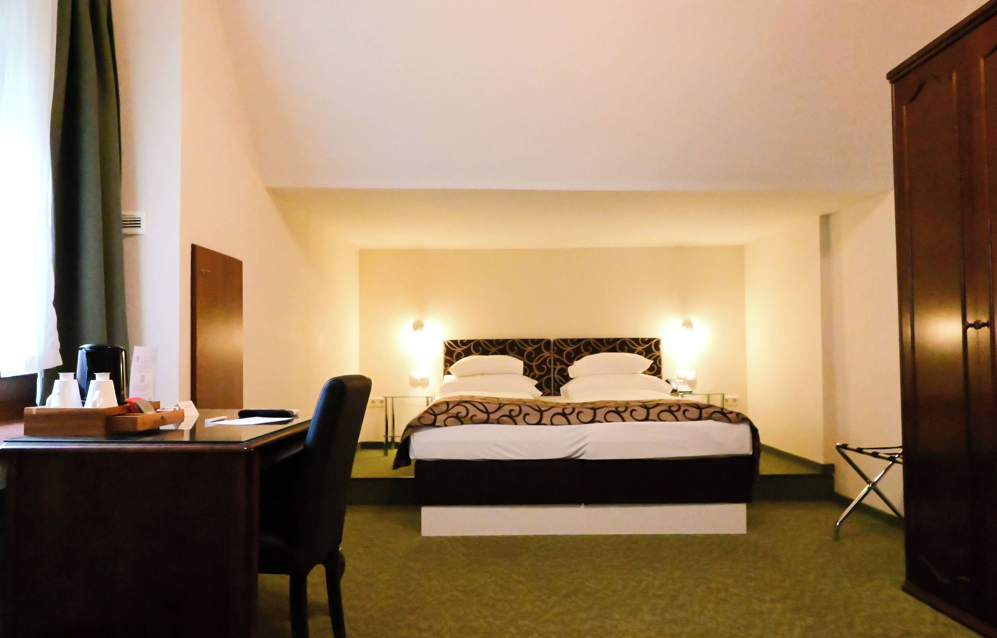 A hotel room with a bed, a desk with a chair, a wardrobe, and a bedside table with a lamp.