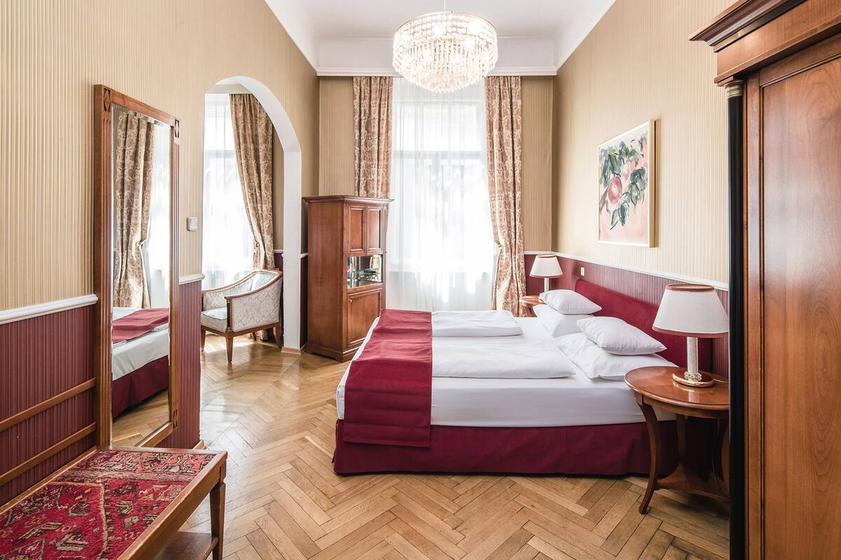 Une chambre d’hôtel avec un grand lit, une armoire en bois, un bureau avec une chaise, un lustre et un tableau au mur.