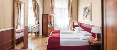 A hotel room with a large bed, a wooden wardrobe, a desk with a chair, a chandelier, and a painting on the wall.