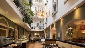 Een hotel lobby met een hoge plafond, een kroonluchter, een glazen vitrine met gebak en een zithoek met tafels en stoelen.