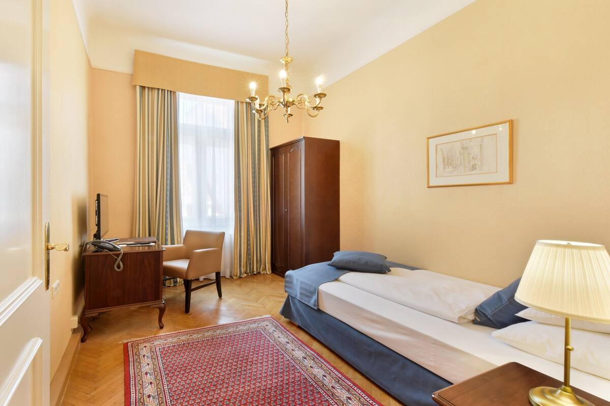 Une chambre d’hôtel comprenant un lit, un bureau, une chaise, une armoire, un lustre, un tableau encadré et un tapis rouge à motifs.