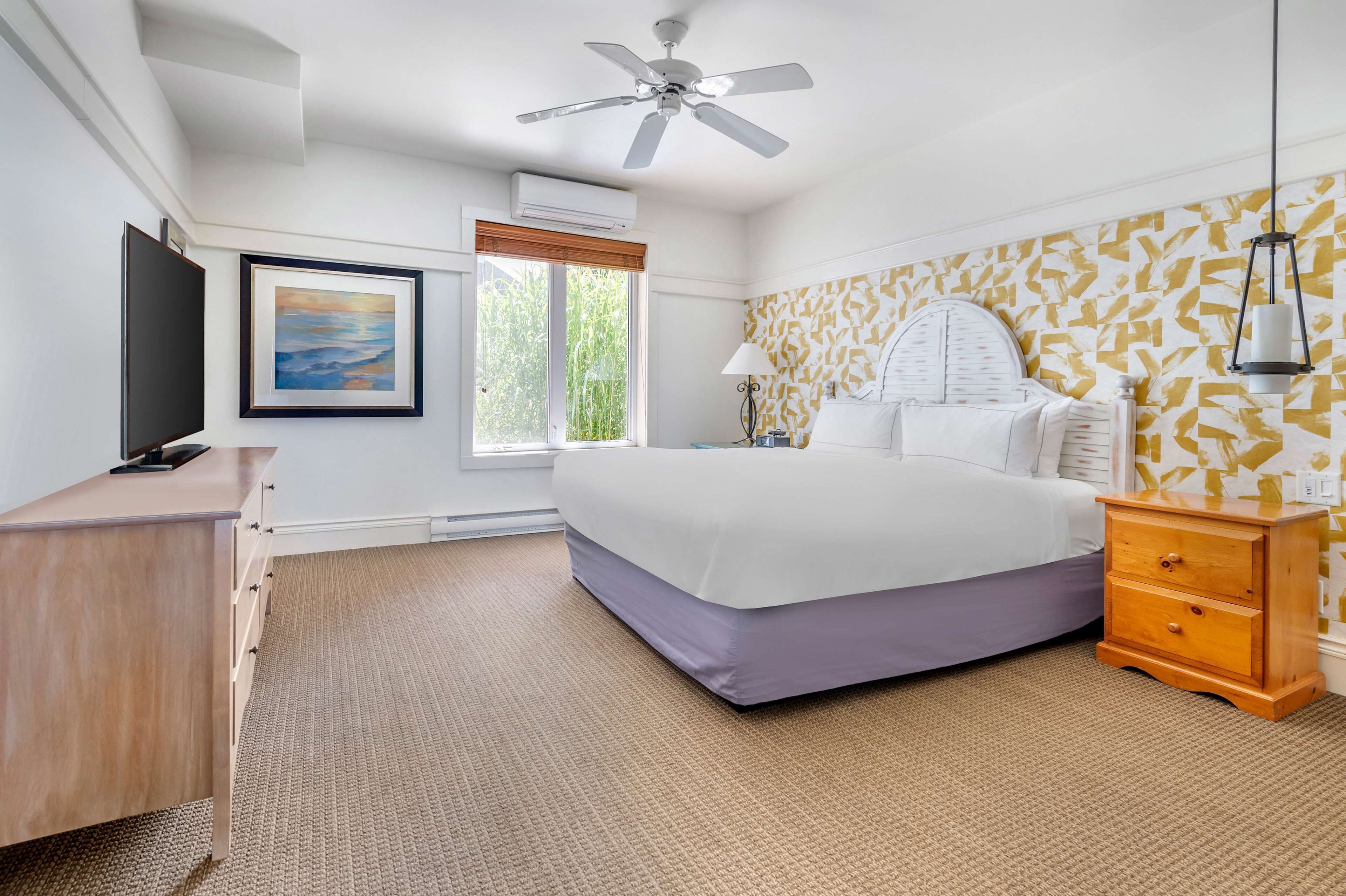 A bedroom with a bed, a desk, a dresser, a ceiling fan, a window with blinds, and a wall with patterned wallpaper.