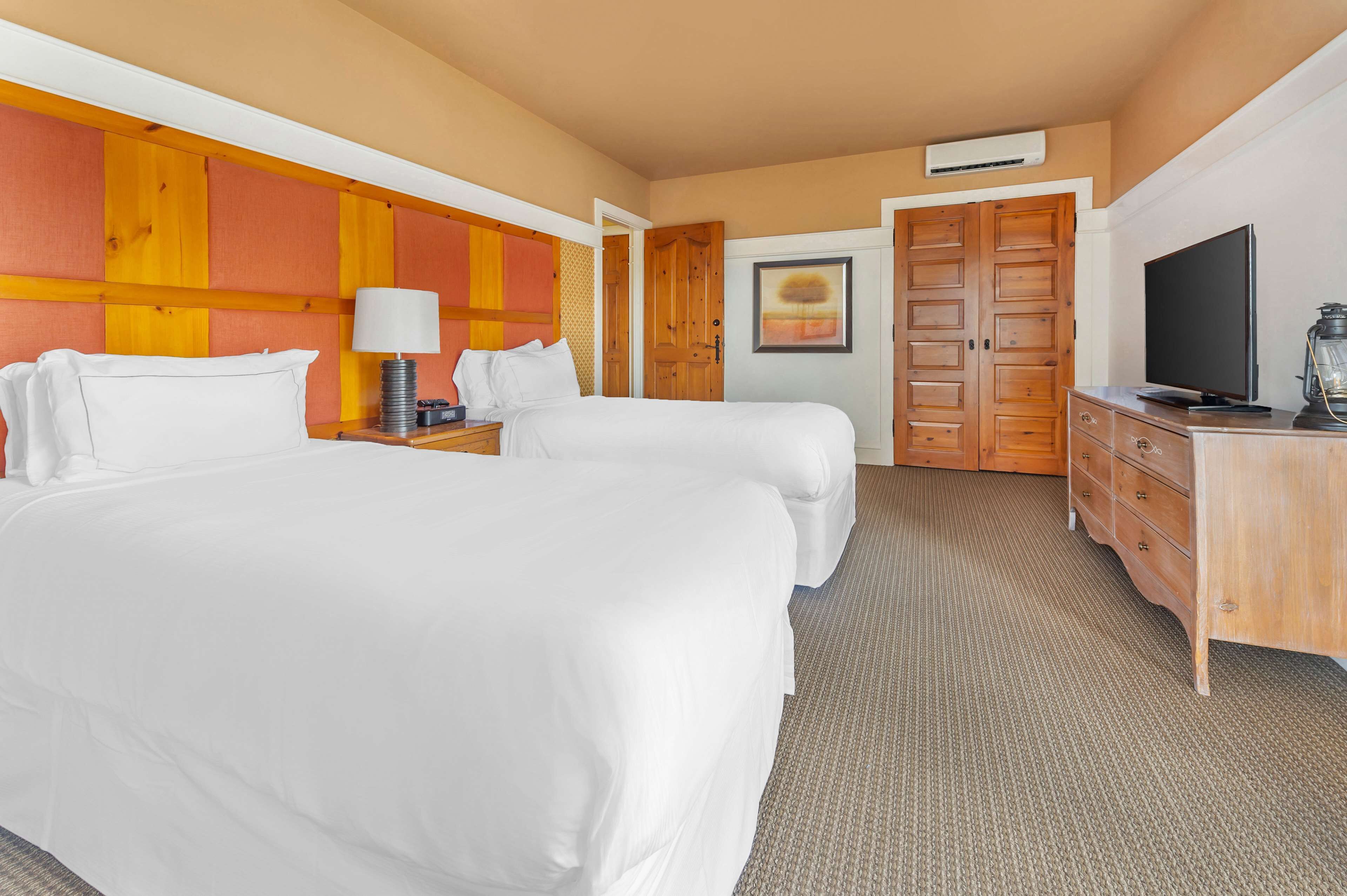 A hotel room with two beds, a wooden headboard, a television, a desk with a lamp, and a wardrobe.