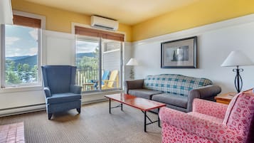 Un salon avec un fauteuil bleu, un canapé gris, une chaise à motifs rouges, une table basse en bois et une vue sur un balcon aménagé avec du mobilier d’extérieur.
