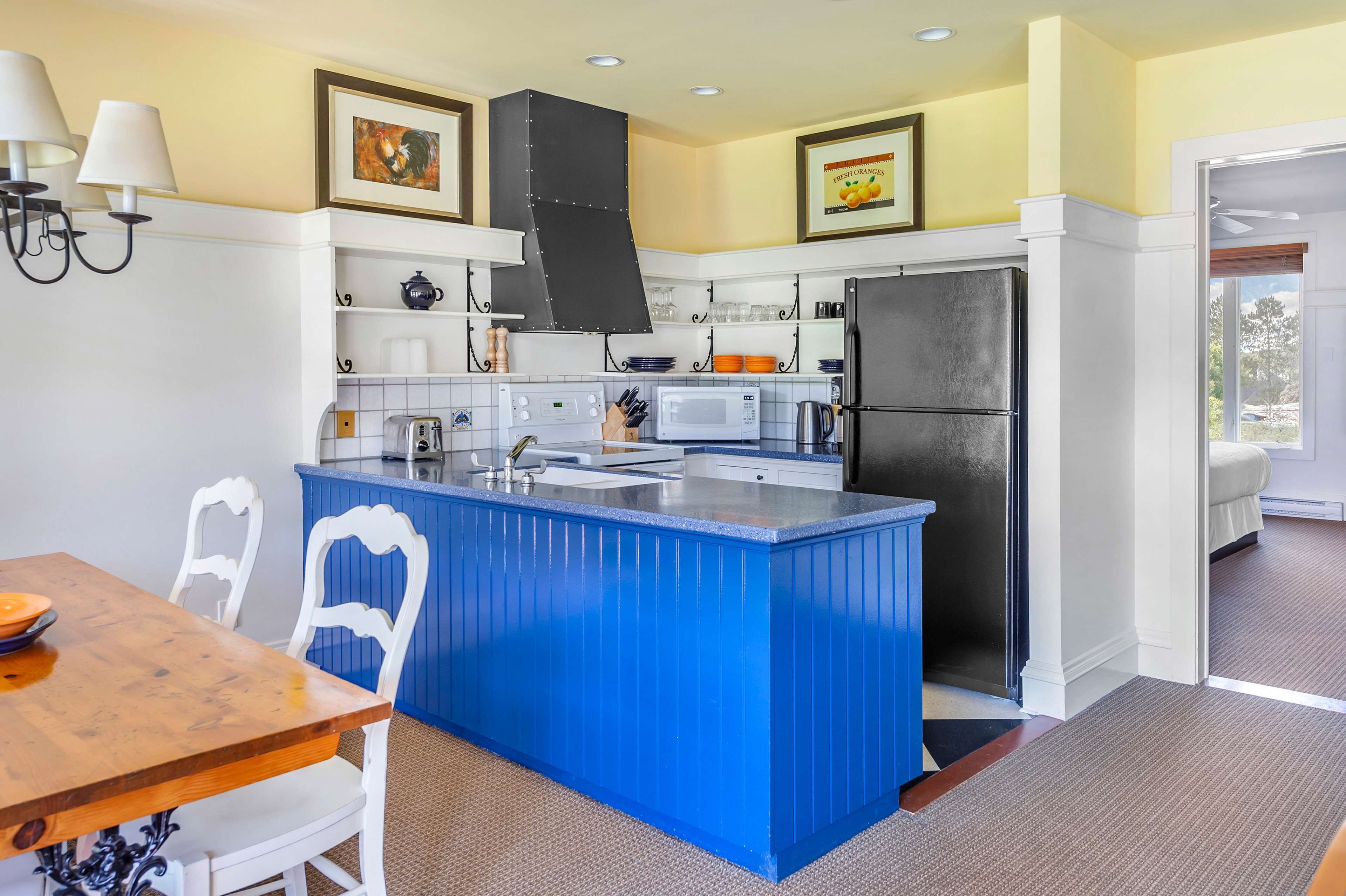 Una cocina con una isla azul, armarios blancos, un refrigerador negro y un comedor con una mesa de madera y sillas blancas.