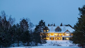 Une grande maison à étages, dont le toit est recouvert de neige et dont le porche avant est bien éclairé.