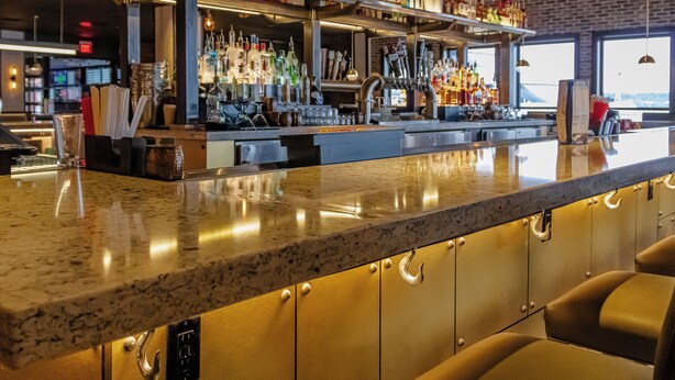 A bar area with a polished granite countertop, a variety of liquor bottles, a television screen, and a view of the outdoors through large windows.