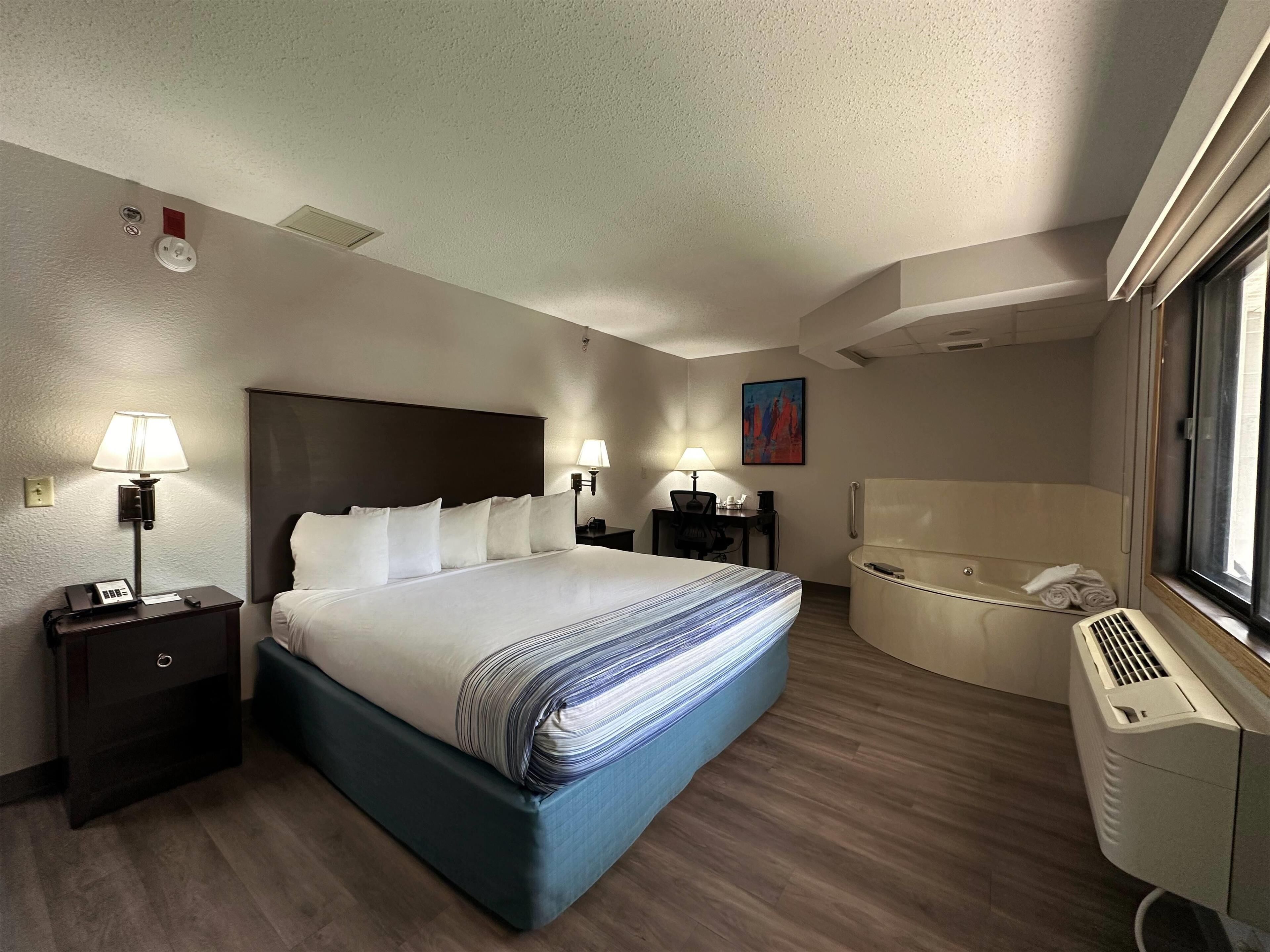 A hotel room with a large bed, bedside lamps, a desk, a chair, a painting, and a separate bathroom with a bathtub.