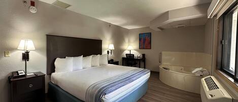 A hotel room with a large bed, bedside lamps, a desk, a chair, a painting, and a separate bathroom with a bathtub.