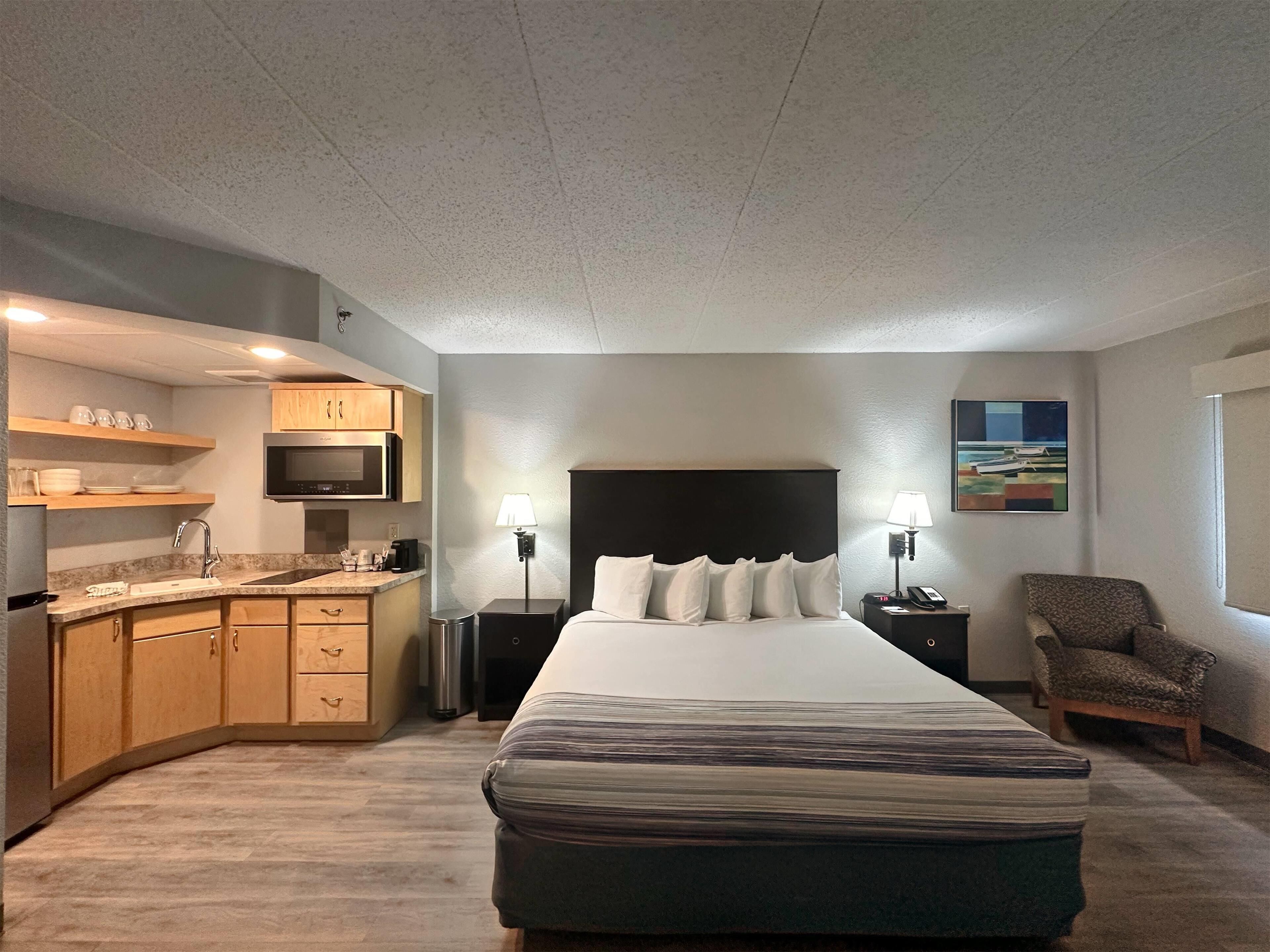 A hotel room with a bed, a desk, a chair, a microwave, a sink, and a painting on the wall.