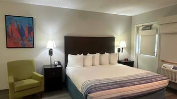 A hotel room with a large bed, two bedside lamps, a nightstand, a green armchair, a painting on the wall, and a door with a mirror.