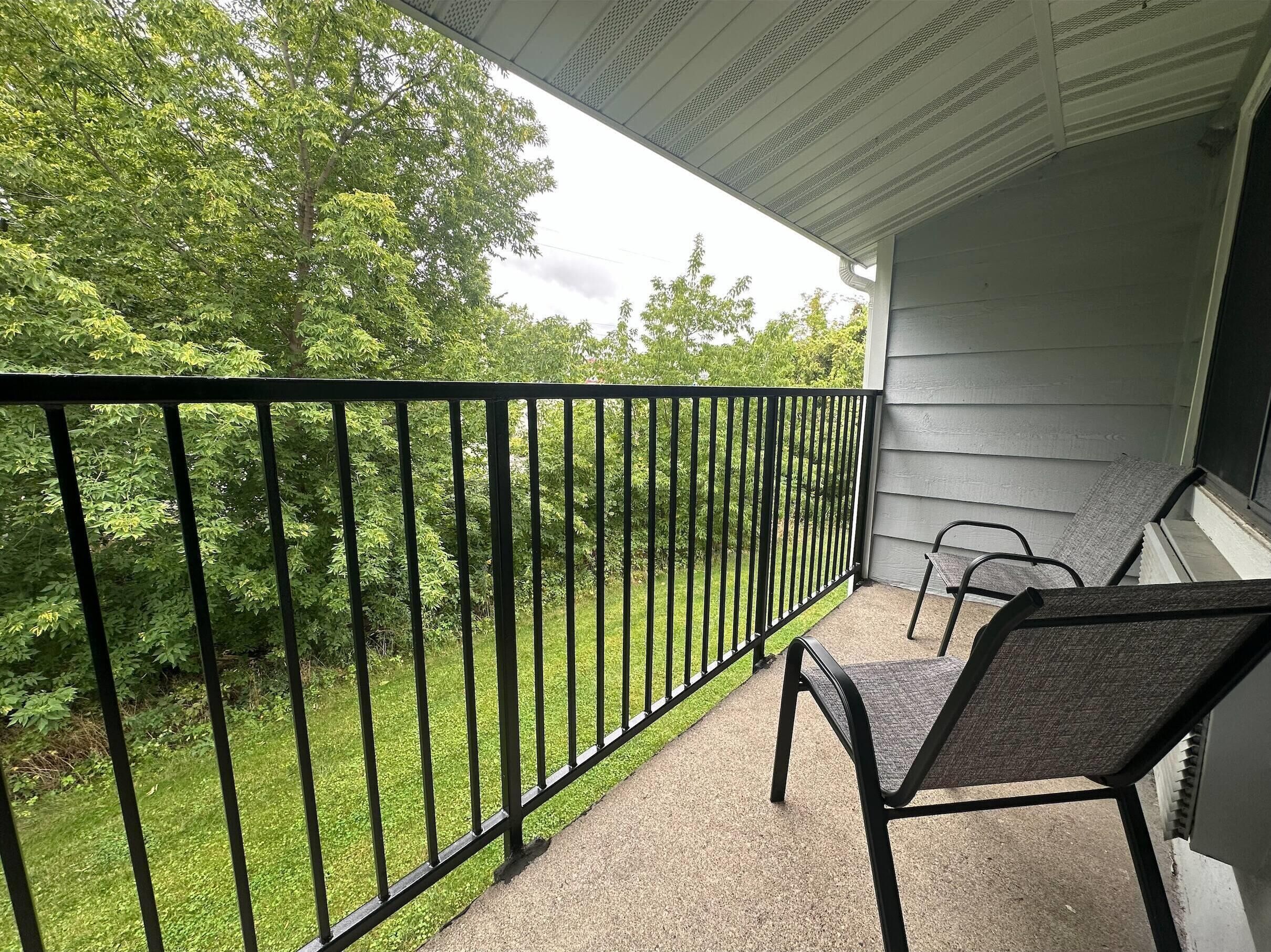 A balcony with a black metal railing, a chair, and a view of a green lawn and trees.