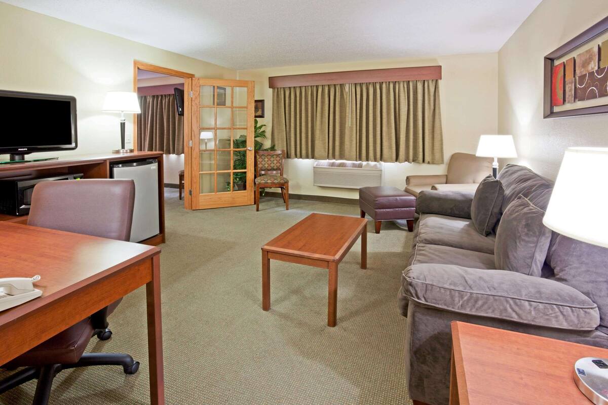 Habitación de hotel con escritorio, sofá, televisor, lámpara y ventana con cortinas.