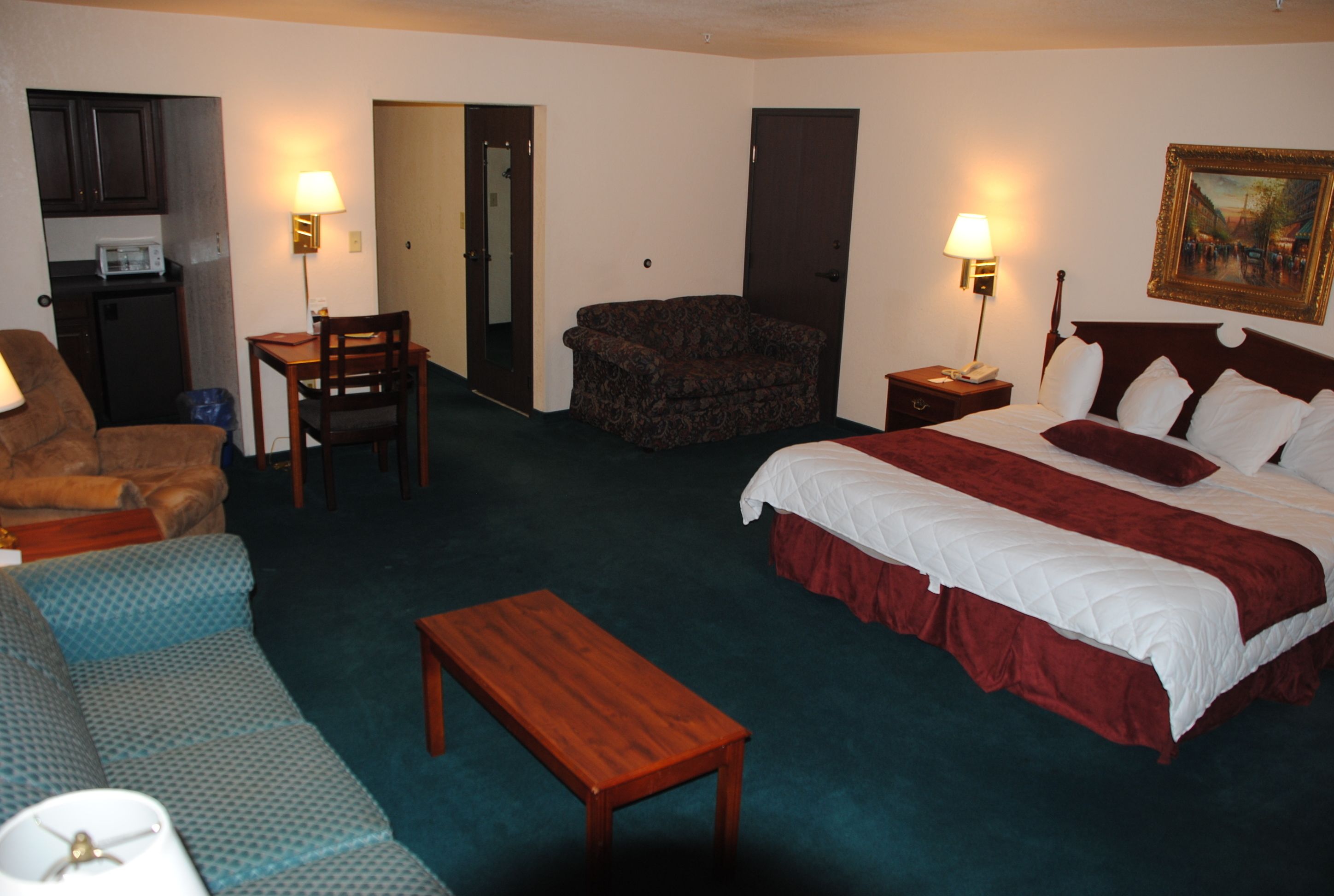 Ein Hotelzimmer mit einem Bett, einem Sofa, einem Sessel, einem kleinen Tisch und einem Schreibtisch. Eine Tür führt zu einer Küchenzeile.