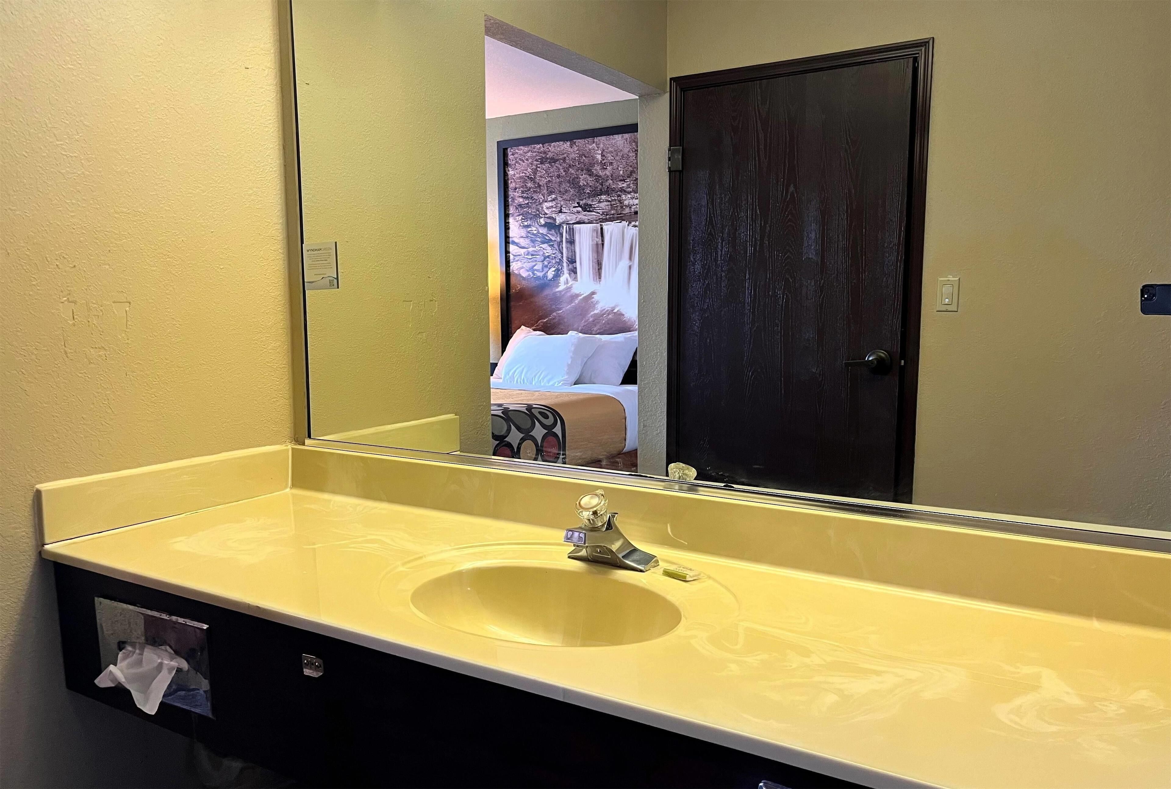 A bathroom with a yellow sink, a dark door, and a mirror reflecting a bed and a waterfall.