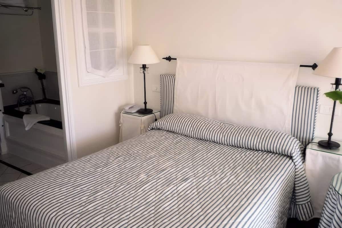 Ein Schlafzimmer mit einem Bett, zwei Nachttischlampen, einem Nachttisch, einem Fenster mit Vorhängen, einem Badezimmer mit Badewanne und einer Topfpflanze.