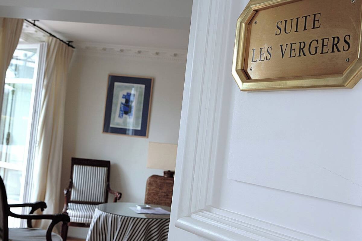 Ein Hotelzimmer mit einem Schild, auf dem 'SUITE LES VERGERS' steht. An der Wand hängt ein gerahmtes Bild, es gibt einen Sessel, einen kleinen Tisch und ein Fenster mit Vorhängen.