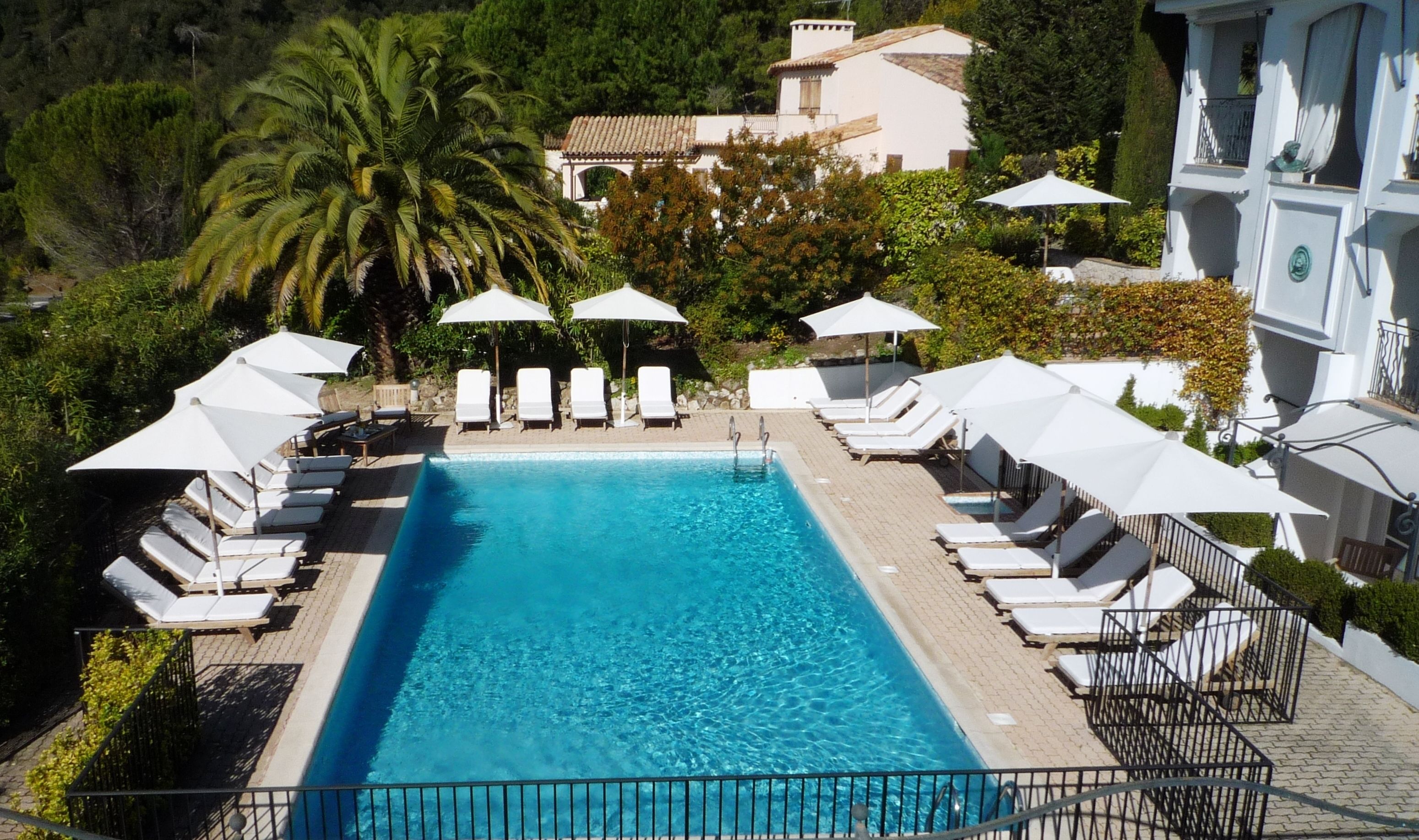 A swimming pool with lounge chairs and umbrellas, surrounded by greenery and a building in the background.