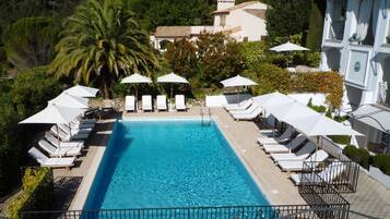 A swimming pool with lounge chairs and umbrellas, surrounded by greenery and a building in the background.