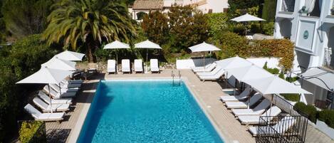 Ein Swimmingpool mit Liegestühlen und Sonnenschirmen, umgeben von viel Grün und einem Gebäude im Hintergrund.
