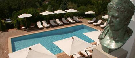 A swimming pool with lounge chairs and umbrellas, surrounded by greenery and a sculpture.