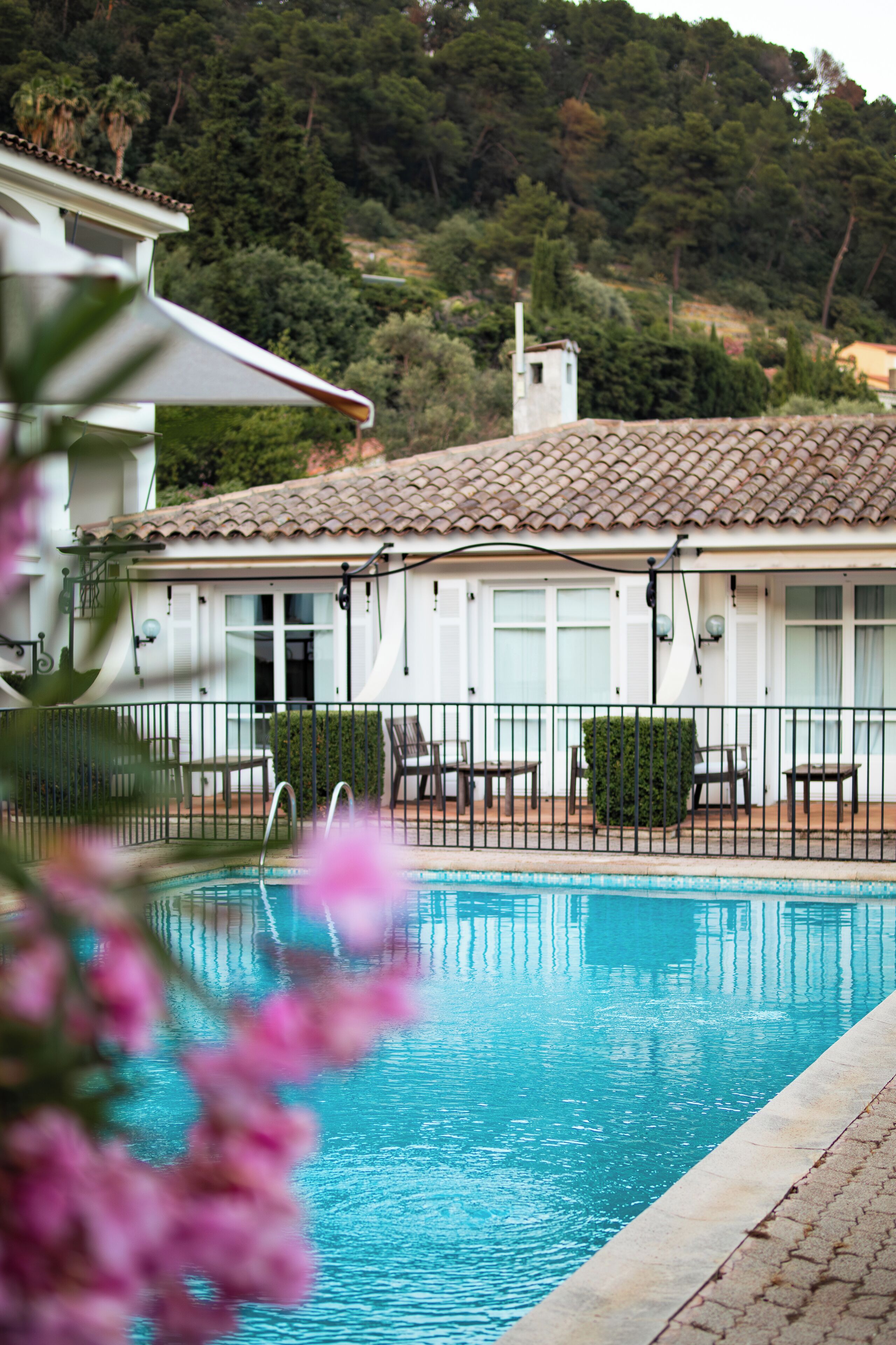Una piscina con borde de baldosas, rodeada por un edificio blanco con tejado de cerámica y un paisaje montañoso de fondo.