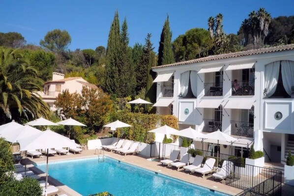 Un área junto a la piscina con sillones de descanso y sombrillas, piscina y un edificio con balcones.