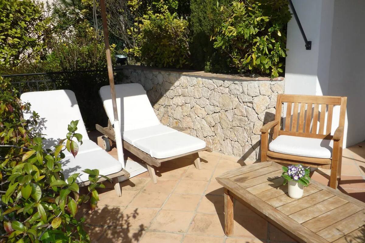 Eine Terrasse mit zwei weißen Liegestühlen, einem Holzstuhl und einem kleinen Tisch mit einem Blumentopf.