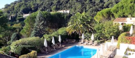 A swimming pool with lounge chairs and umbrellas, surrounded by lush greenery and a hillside.