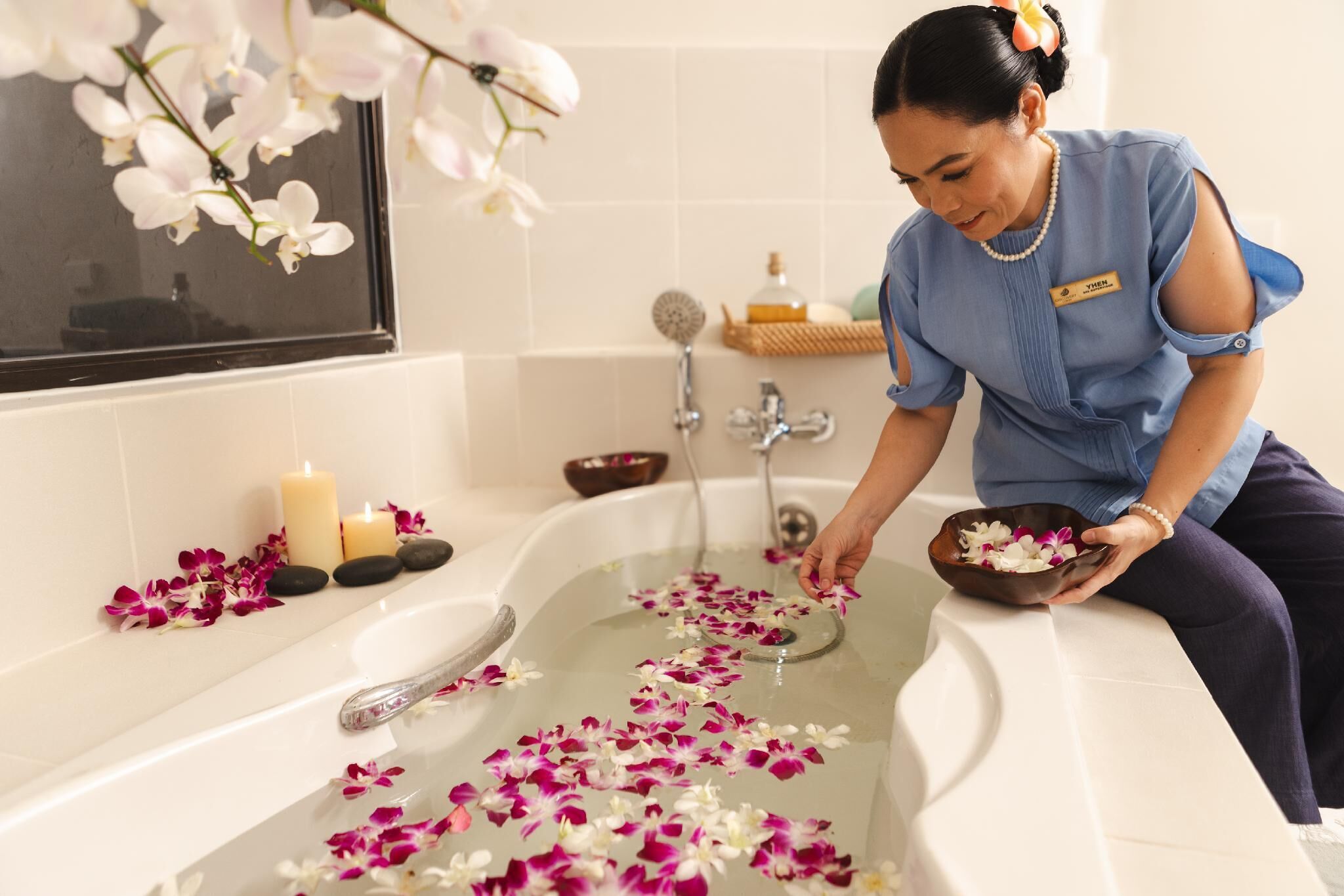 Une personne vêtue d’un uniforme bleu prépare un bain agrémenté de pétales de rose et de bougies, dans une salle de bain équipée d’une baignoire, d’une fenêtre et d’une étagère contenant des articles de toilette.