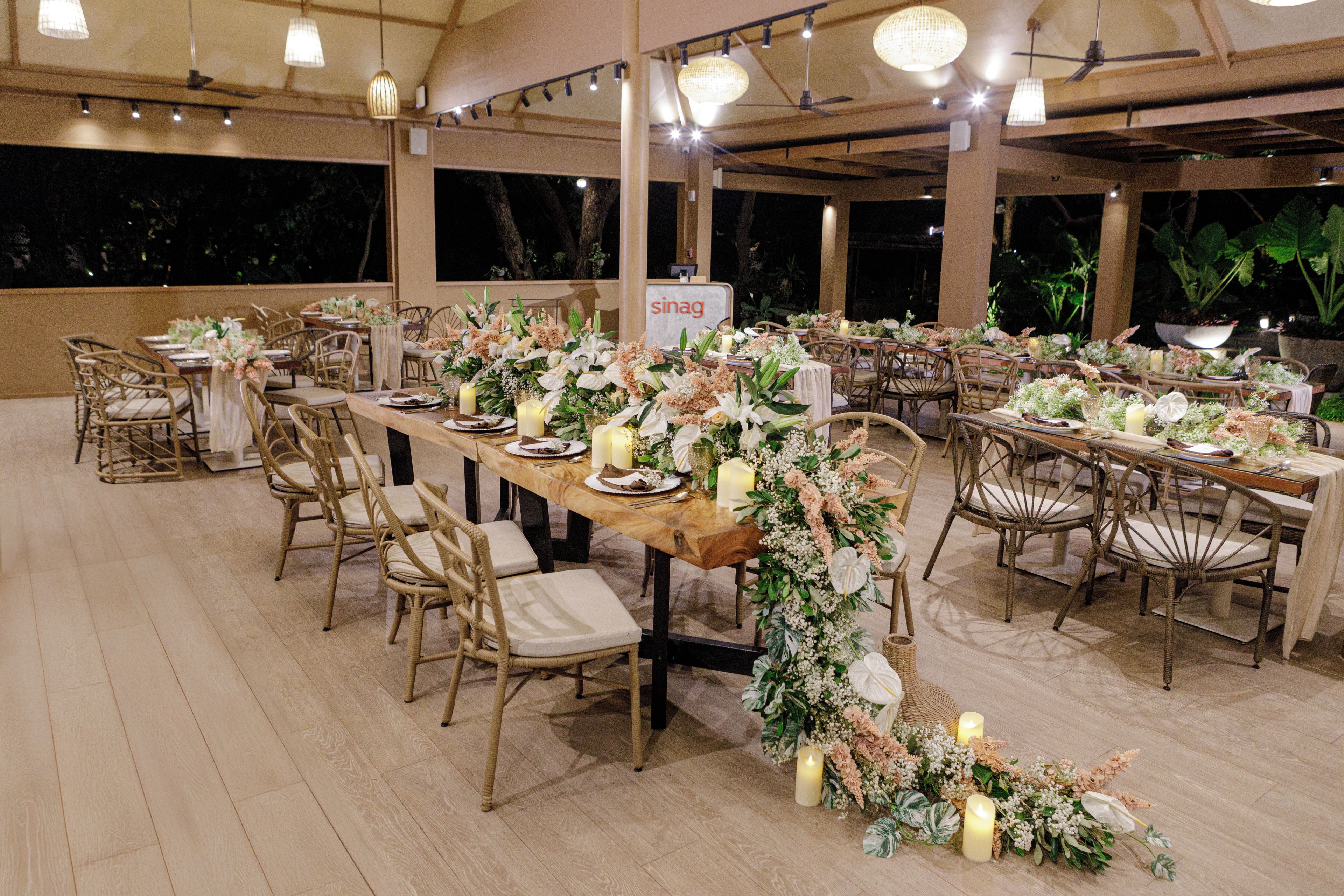 An outdoor dining area with multiple tables set for a formal event, featuring floral centerpieces and candles.