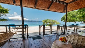 Terraza de madera con mobiliario de mimbre, con vistas a una playa de aguas azul turquesa y montañas a lo lejos.
