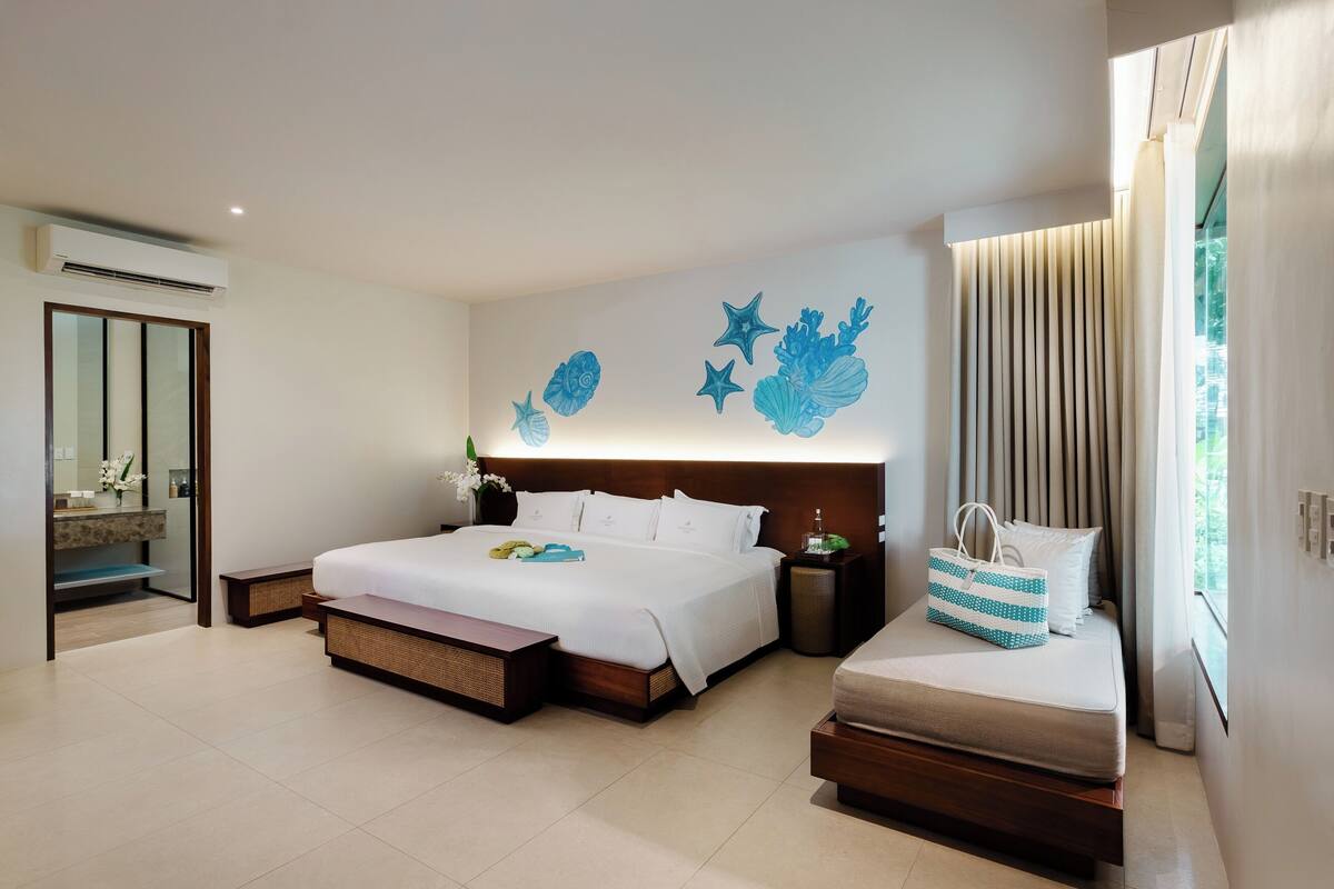Quarto de hotel moderno com uma cama grande, mesas de cabeceira, um sofá pequeno, um mural decorativo na parede e uma janela ampla com cortinas.