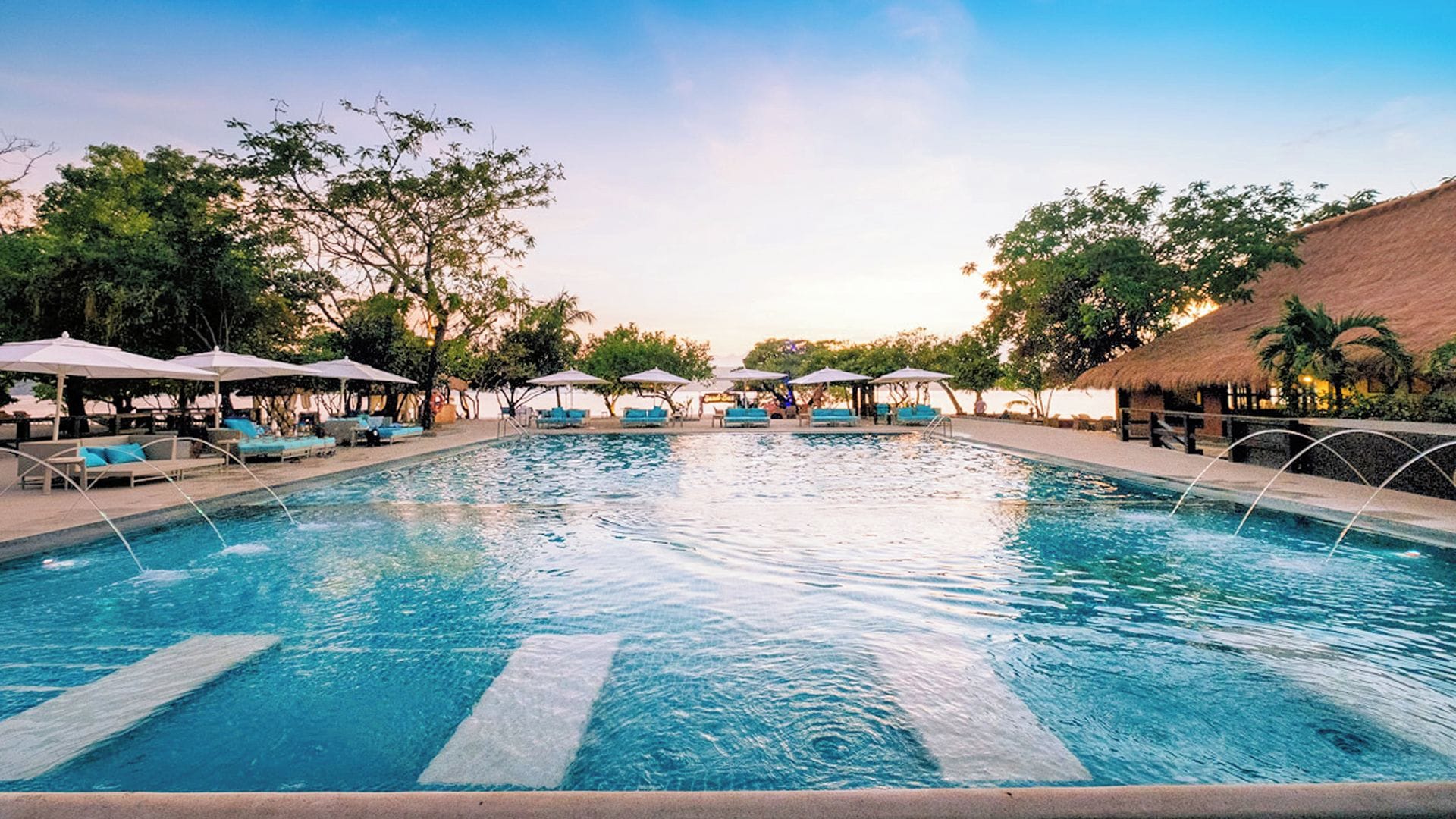 A swimming pool with lounge chairs and umbrellas, surrounded by trees and a thatched-roof structure.