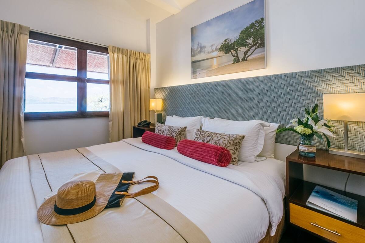 Cama bem arrumada com chapéu e bolsa em cima, mesa de cabeceira com luminária e quadro na parede.