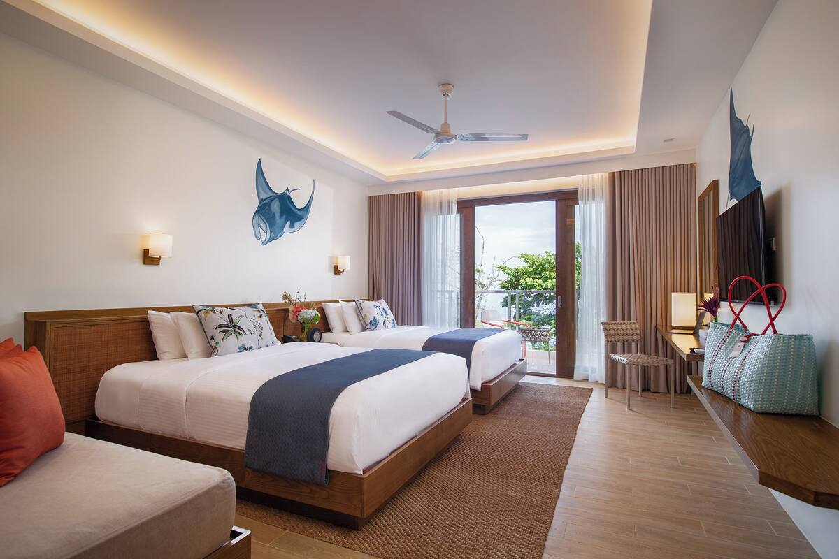 Quarto de hotel com duas camas, uma escrivaninha, uma cadeira, televisão e varanda com cortinas.