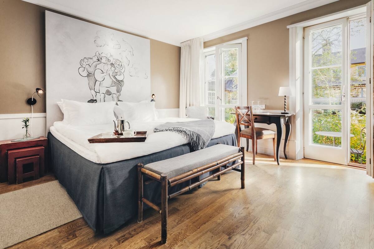 Un dormitorio con una cama grande, mesitas de noche, un banco, un escritorio y vistas al exterior.