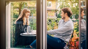 Una pareja sentada a una mesa con un calentador de agua y tazas, en una habitación con ventanas amplias y vistas a la vegetación.