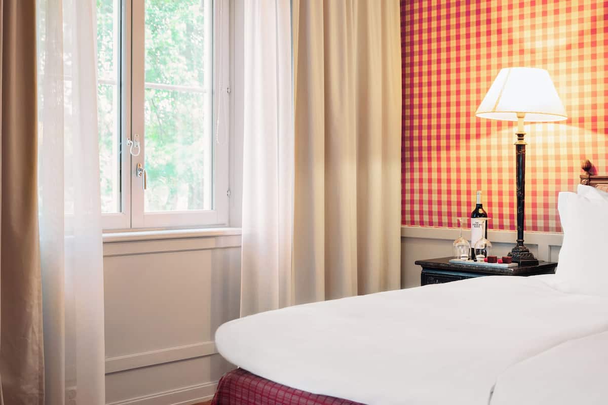 Habitación de hotel con una cama grande, una ventana con cortinas, una mesita de noche con lámpara y un papel tapiz a cuadros.