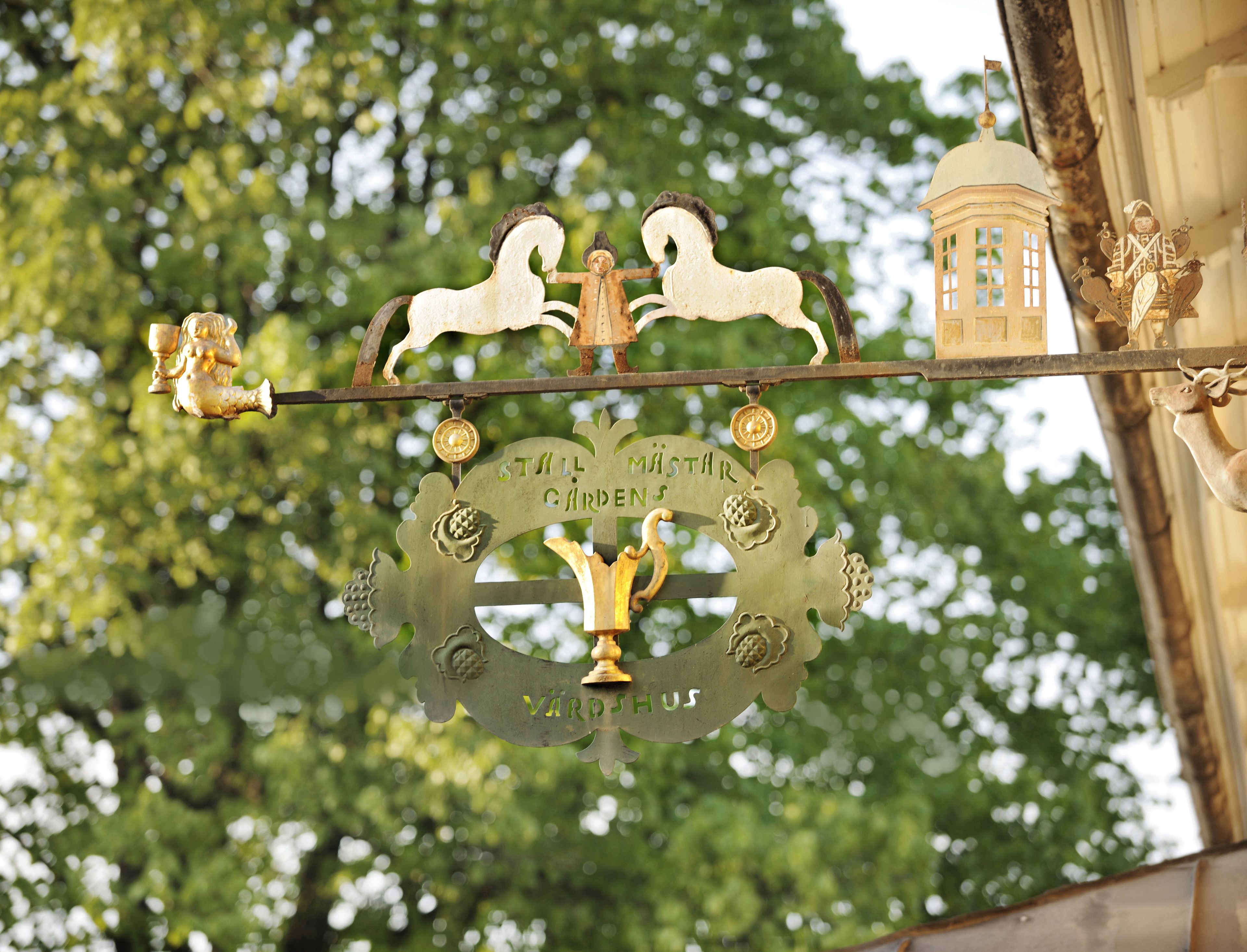 Un letrero de metal decorativo con un jinete a caballo, y el texto "STILL MÄSTAR GARDENS" y "VÄRD SÖR".