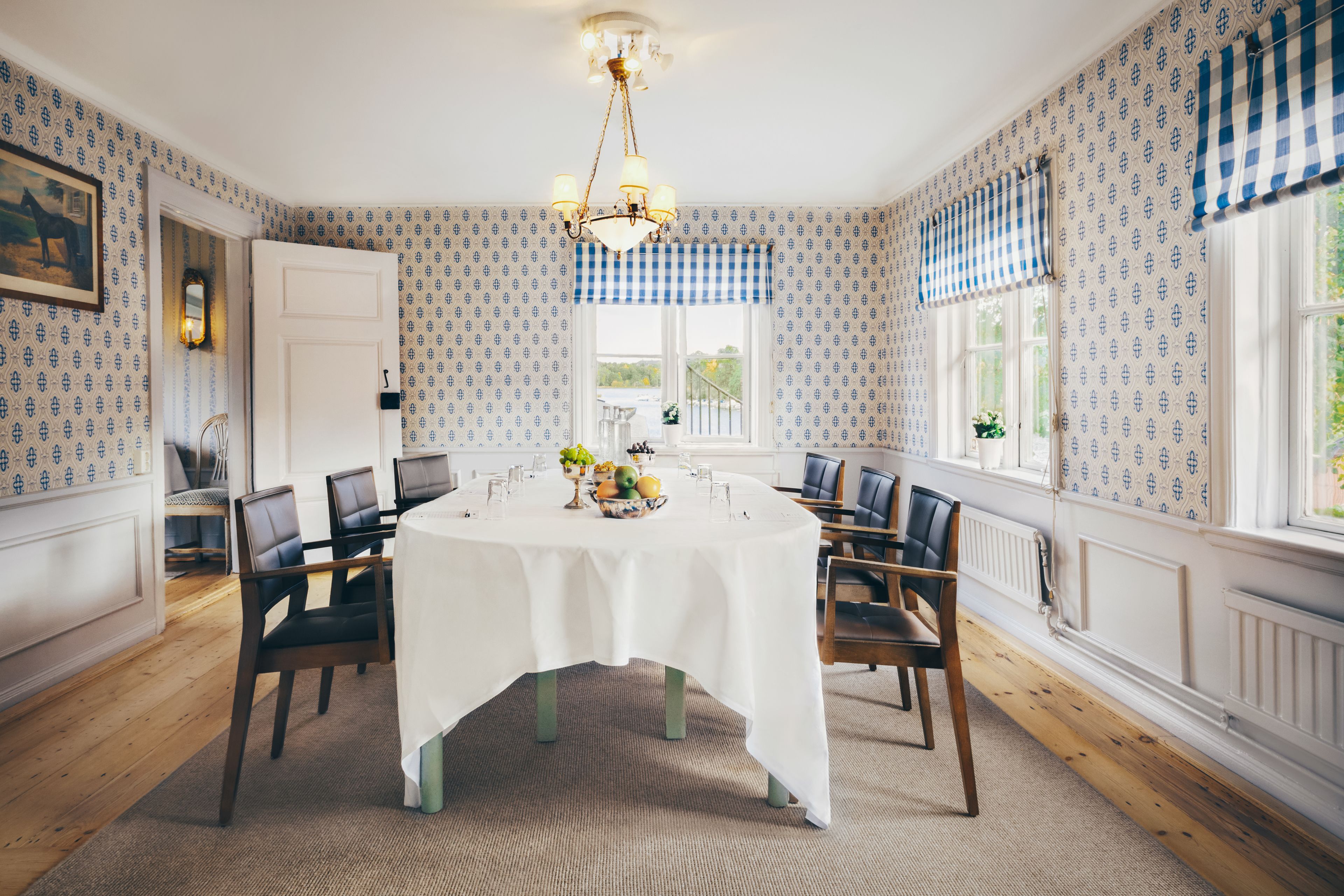 Uma sala de jantar com uma mesa redonda, cadeiras e um candelabro. Há janelas com cortinas quadriculadas azuis e brancas, e uma porta que dá para outro cômodo.