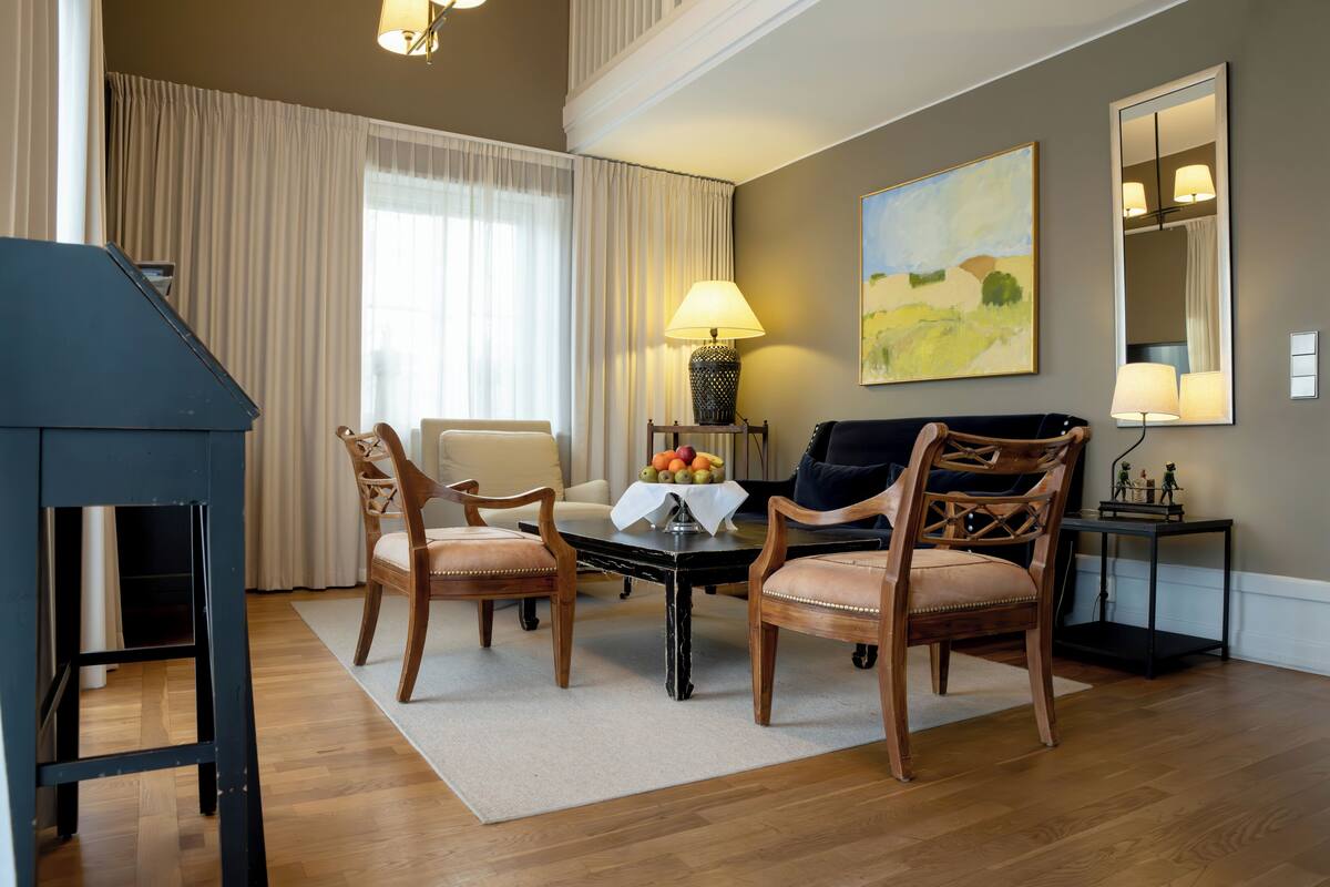 Una sala de estar con chimenea, dos sillas de madera, una mesa con fruta, una lámpara y un cuadro en la pared.