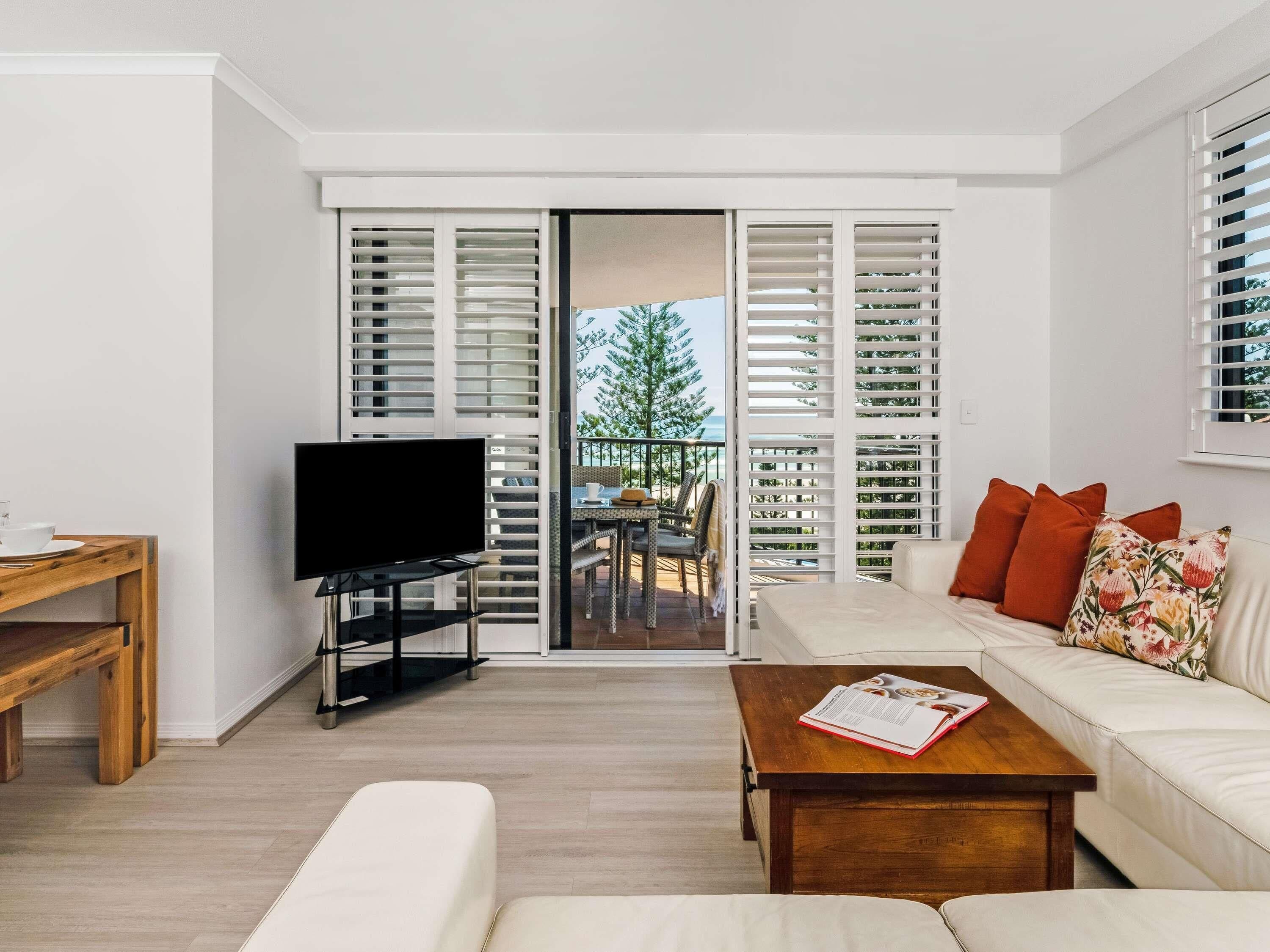 A modern living room with a flat-screen TV, a sofa with cushions, a wooden coffee table, and a sliding glass door leading to an outdoor area with a view of a tree and a building.