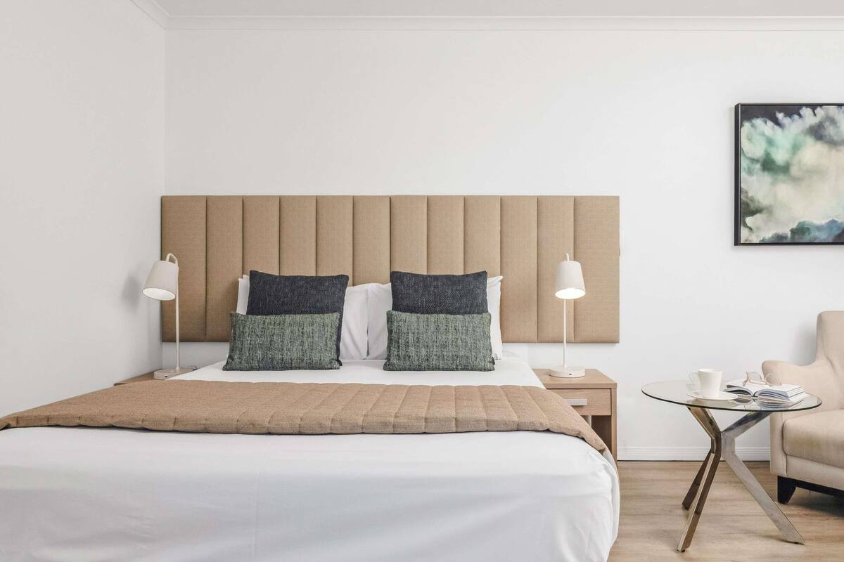 A modern hotel room with a large bed, two bedside lamps, a small side table, and a chair.