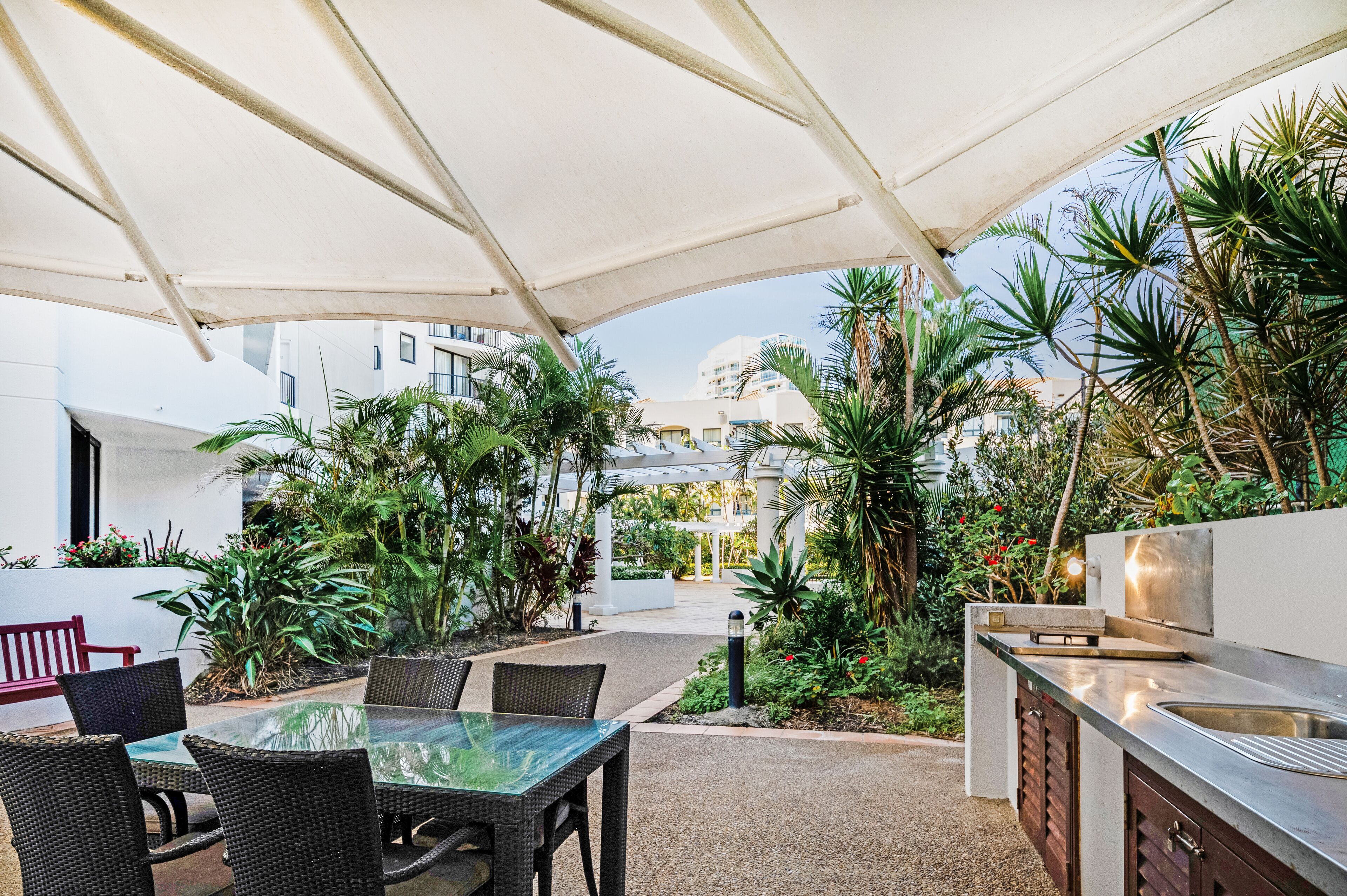 An outdoor patio area with a glass table and chairs, surrounded by lush greenery and a white canopy.