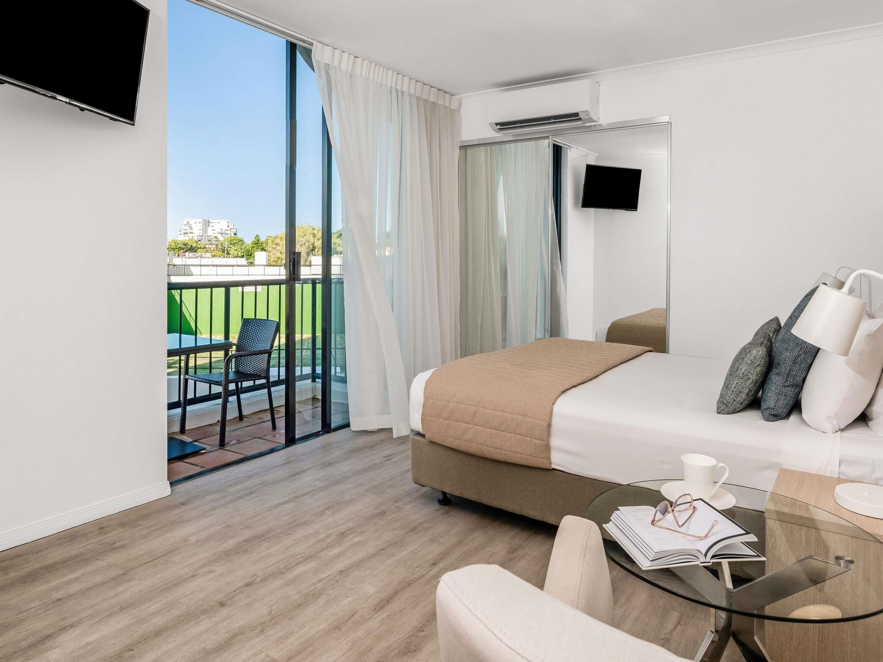A hotel room with a bed, a glass table with a book and glasses, a TV, and a balcony with a chair and table.